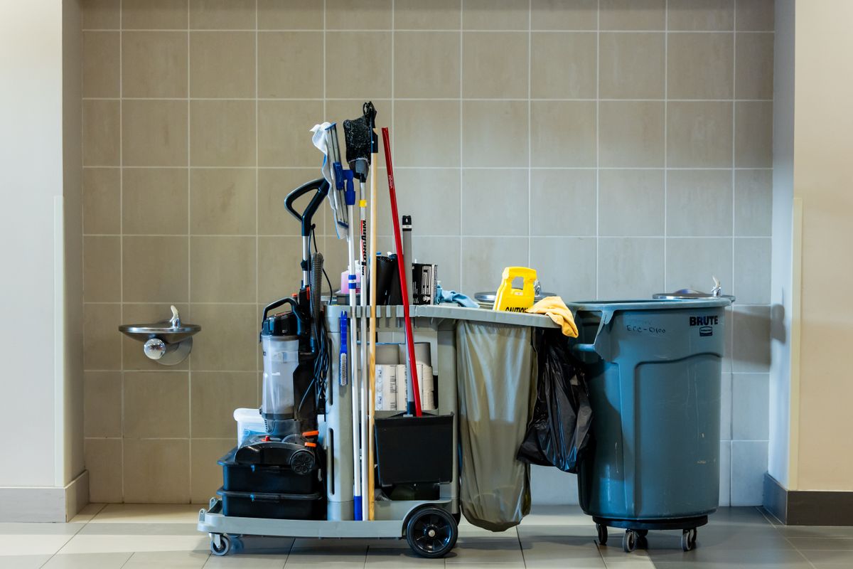 Janitorial cart with mops, a vacuum, paper supplies and a caution sign in a tiled commercial corridor