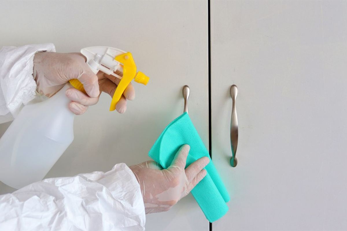 Gloved hands disinfecting cabinet door handles with a spray bottle and cloth